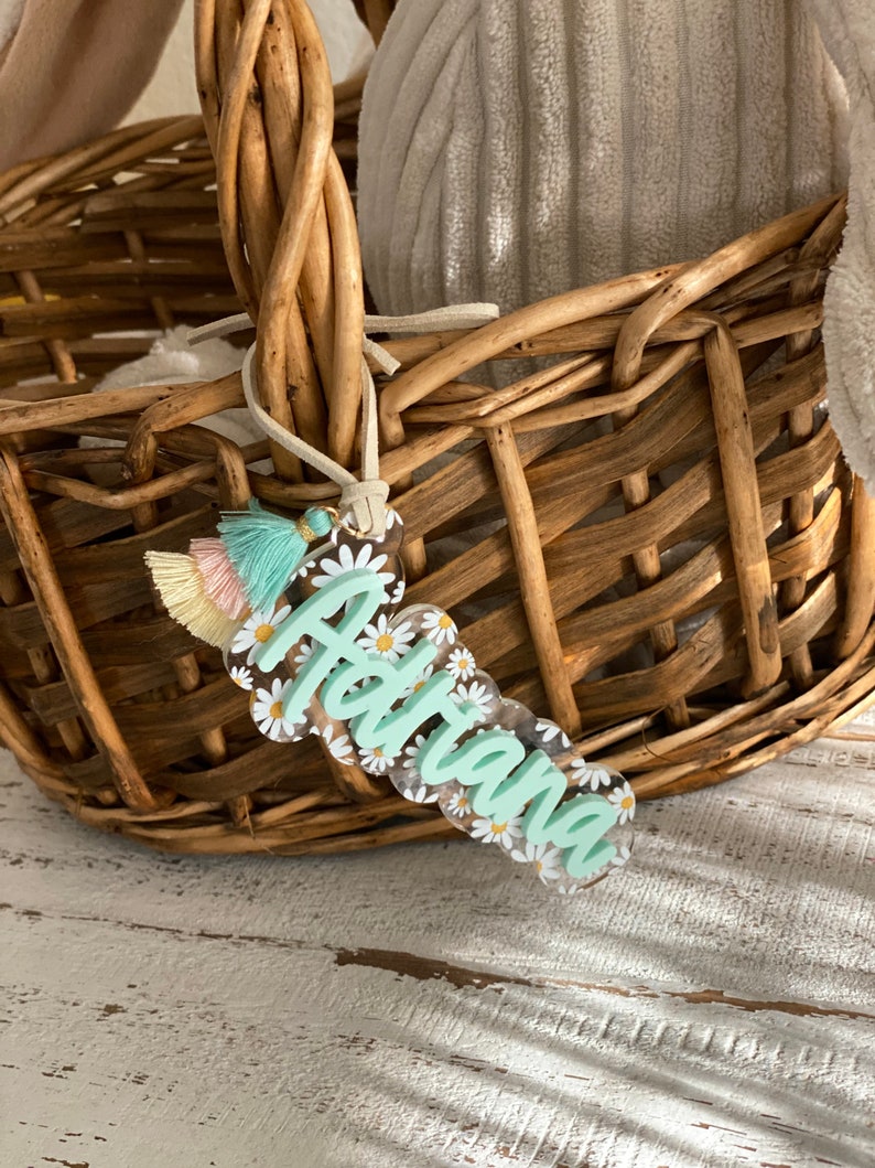 May include: A clear acrylic name tag with the name "Adriana" in a light green script, surrounded by white daisies. The tag has a scalloped edge and a tassel with pastel colors. The tag is hanging from a woven basket.