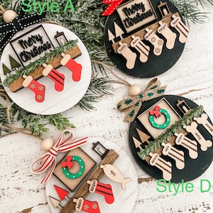 May include: Four different styles of Christmas ornaments featuring a wooden base with a "Merry Christmas" sign, a fireplace, and stockings with personalized names. The ornaments are decorated with greenery, a wreath, and a lantern.