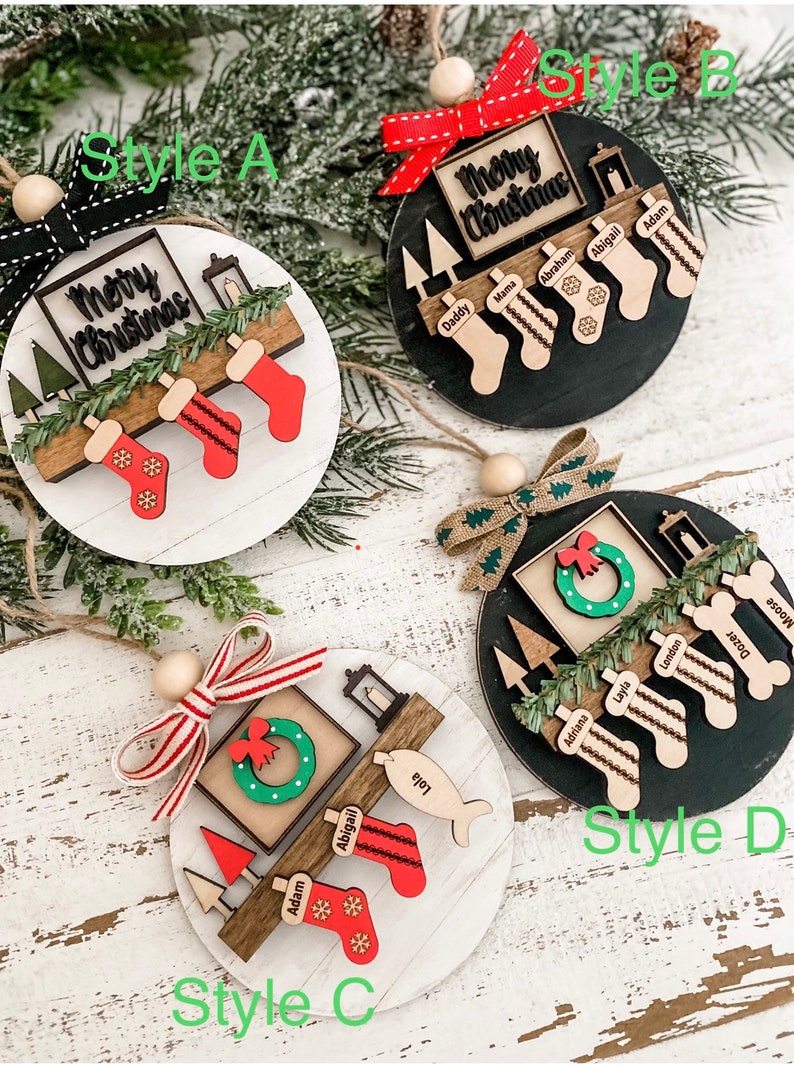 May include: Four different styles of Christmas ornaments featuring a wooden base with a "Merry Christmas" sign, a fireplace, and stockings with personalized names. The ornaments are decorated with greenery, a wreath, and a lantern.