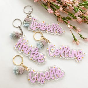 May include: Keychains with names in a light purple script font. Each keychain has a daisy pattern background and a tassel in shades of pink, blue, and tan. The keychains are attached to a silver or gold keyring.