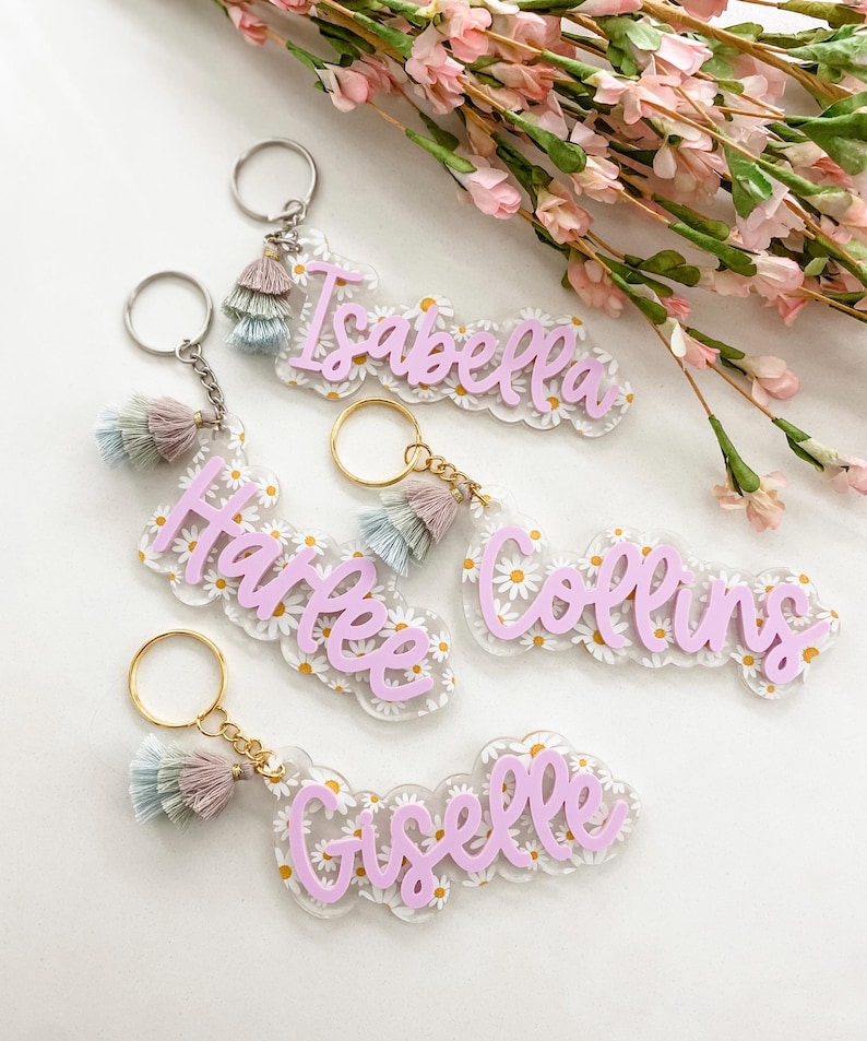 May include: Keychains with names in a light purple script font. Each keychain has a daisy pattern background and a tassel in shades of pink, blue, and tan. The keychains are attached to a silver or gold keyring.
