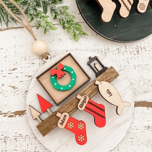 May include: A white wooden Christmas ornament with a red bow, a wreath, a candle, a fish, and stockings with the names Adam, Abigail, and Lola. The ornament is decorated with snowflakes.
