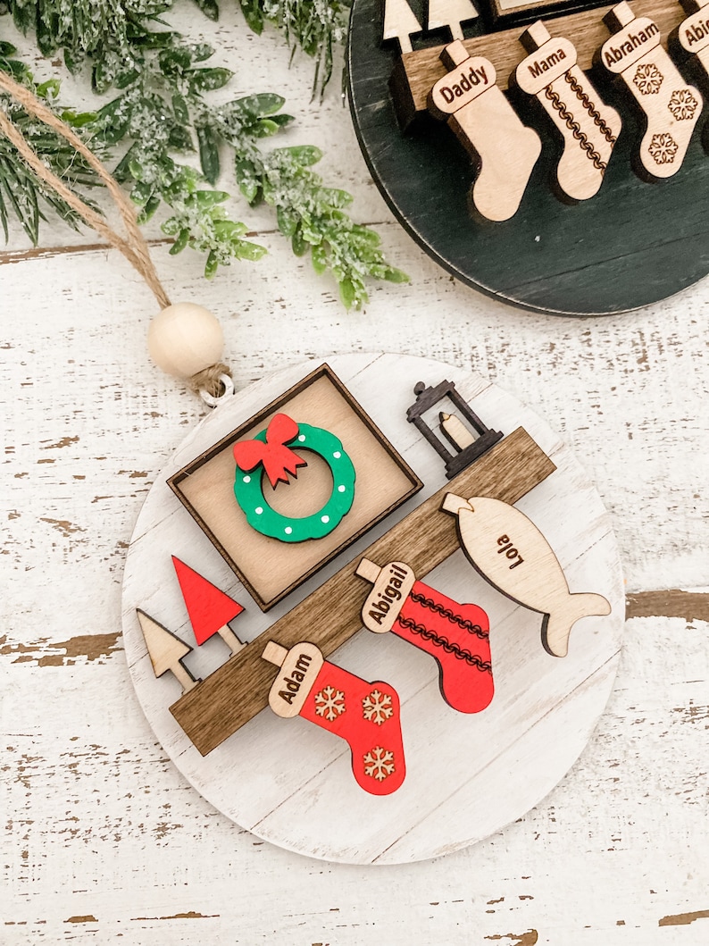 May include: A white wooden Christmas ornament with a red bow, a wreath, a candle, a fish, and stockings with the names Adam, Abigail, and Lola. The ornament is decorated with snowflakes.