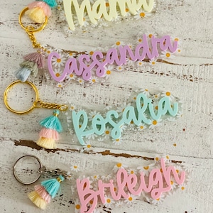 May include: Four personalized keychains with gold rings and colorful tassels. Each keychain features a name in a different pastel color: "Mama", "Cassandra", "Rosalia", and "Brileigh". The background is a clear acrylic with a daisy pattern.