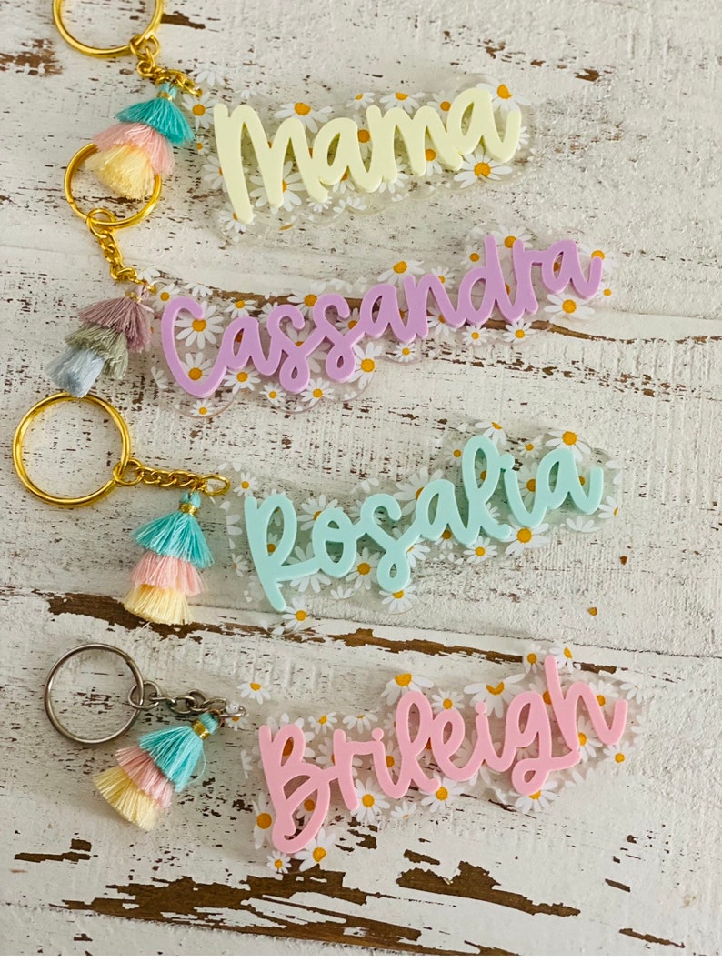 May include: Four personalized keychains with gold rings and colorful tassels. Each keychain features a name in a different pastel color: "Mama", "Cassandra", "Rosalia", and "Brileigh". The background is a clear acrylic with a daisy pattern.