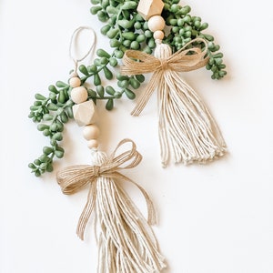 May include: Two handmade Christmas decorations with a rustic, farmhouse style. Each decoration features a wooden bead garland with a geometric wooden bead, a small sprig of faux greenery, and a beige tassel with a burlap bow.