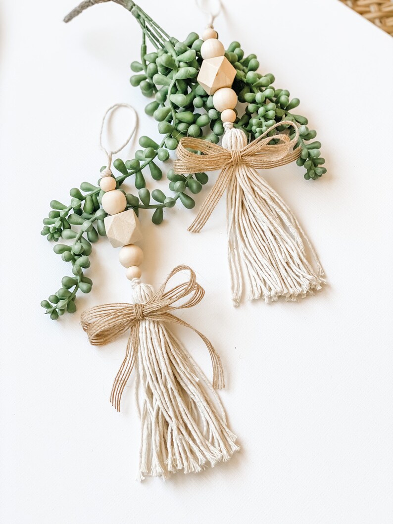 May include: Two handmade Christmas decorations with a rustic, farmhouse style. Each decoration features a wooden bead garland with a geometric wooden bead, a small sprig of faux greenery, and a beige tassel with a burlap bow.