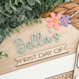 May include: A semi-circular wooden sign featuring the name Bella in raised sage green script. Below the name, a banner reads FIRST DAY OF in black text. Two flowers, one peach and one purple, are attached to the top right corner.