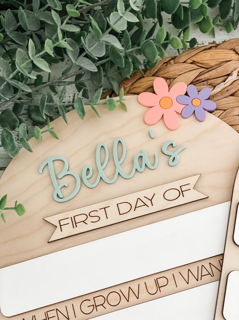 May include: A semi-circular wooden sign featuring the name Bella in raised sage green script. Below the name, a banner reads FIRST DAY OF in black text. Two flowers, one peach and one purple, are attached to the top right corner.