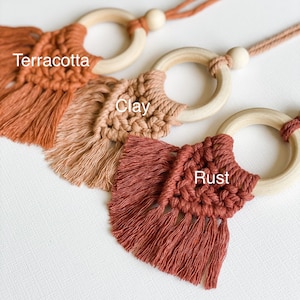May include: Close-up of three macrame car air fresheners in terracotta, clay, and rust colors. Each features a wooden ring and a knotted design with fringe. The text "Terracotta", "Clay", and "Rust" are visible.