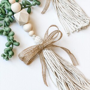 May include: Two white and brown tassel ornaments with a hessian bow. The tassels are made of white string and the bows are made of brown hessian. The ornaments are hanging from a green succulent plant.