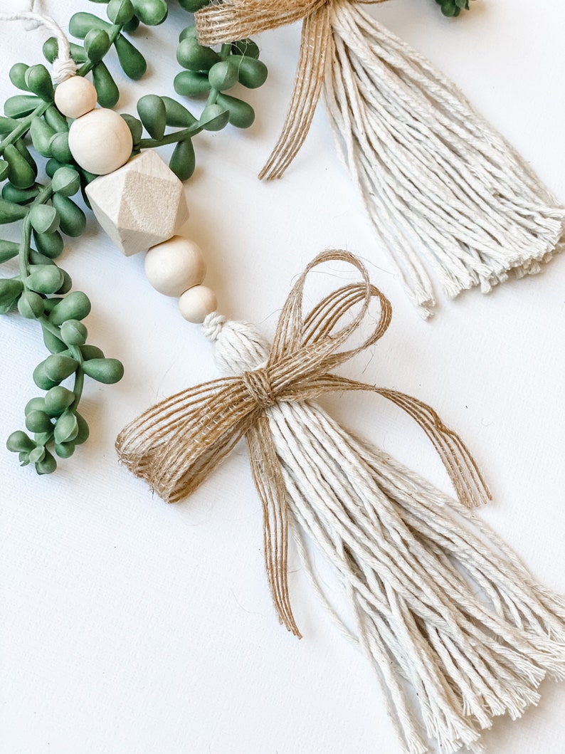 May include: Two white and brown tassel ornaments with a hessian bow. The tassels are made of white string and the bows are made of brown hessian. The ornaments are hanging from a green succulent plant.
