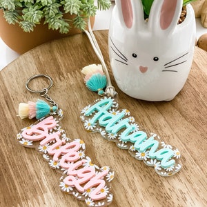 May include: Two personalized acrylic keychains with daisy flower accents. One keychain has the name "Brieley" in pink, the other has the name "Adriana" in light blue. Both keychains have a tassel and a keyring.