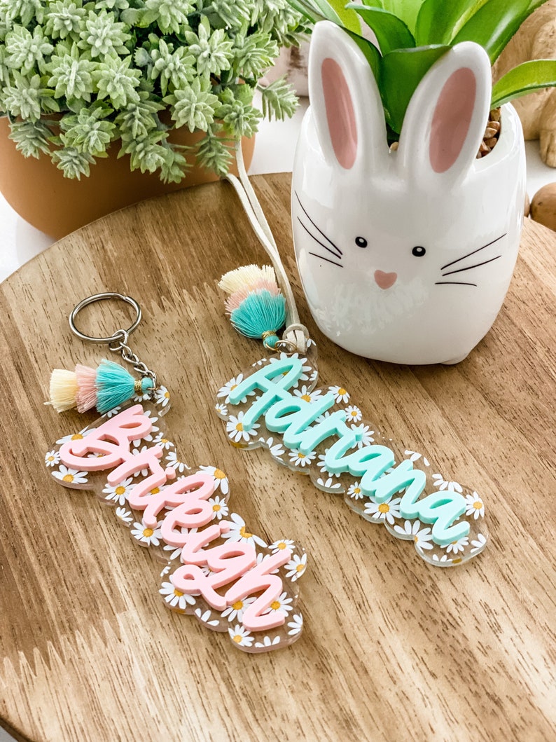 May include: Two personalized acrylic keychains with daisy flower accents. One keychain has the name "Brieley" in pink, the other has the name "Adriana" in light blue. Both keychains have a tassel and a keyring.