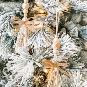 May include: Two beige tassel ornaments with wooden beads and a burlap bow hanging on a snowy Christmas tree.