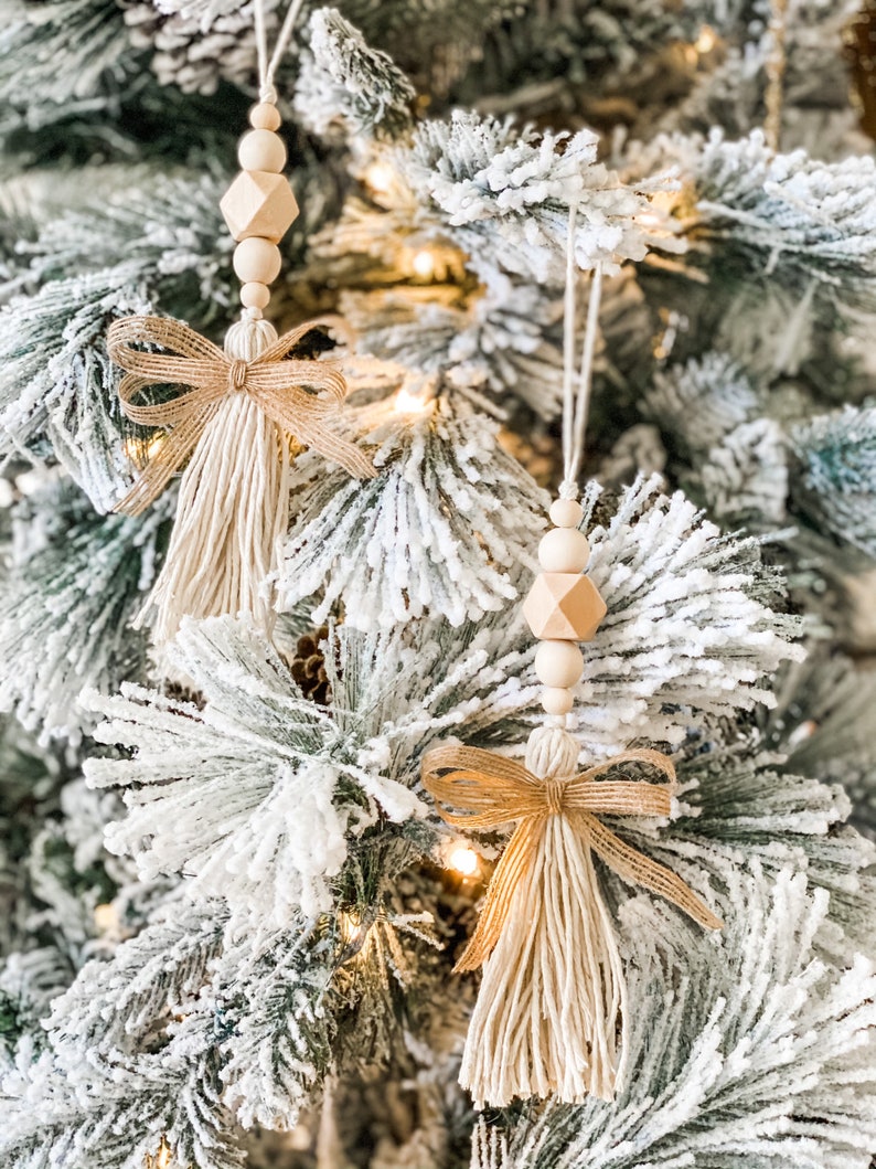 May include: Two beige tassel ornaments with wooden beads and a burlap bow hanging on a snowy Christmas tree.