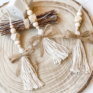 May include: Three wooden bead and tassel ornaments with a natural wood and cream colour scheme. The tassels are made of cream coloured yarn and the beads are natural wood. The ornaments are hanging from a wooden slice.