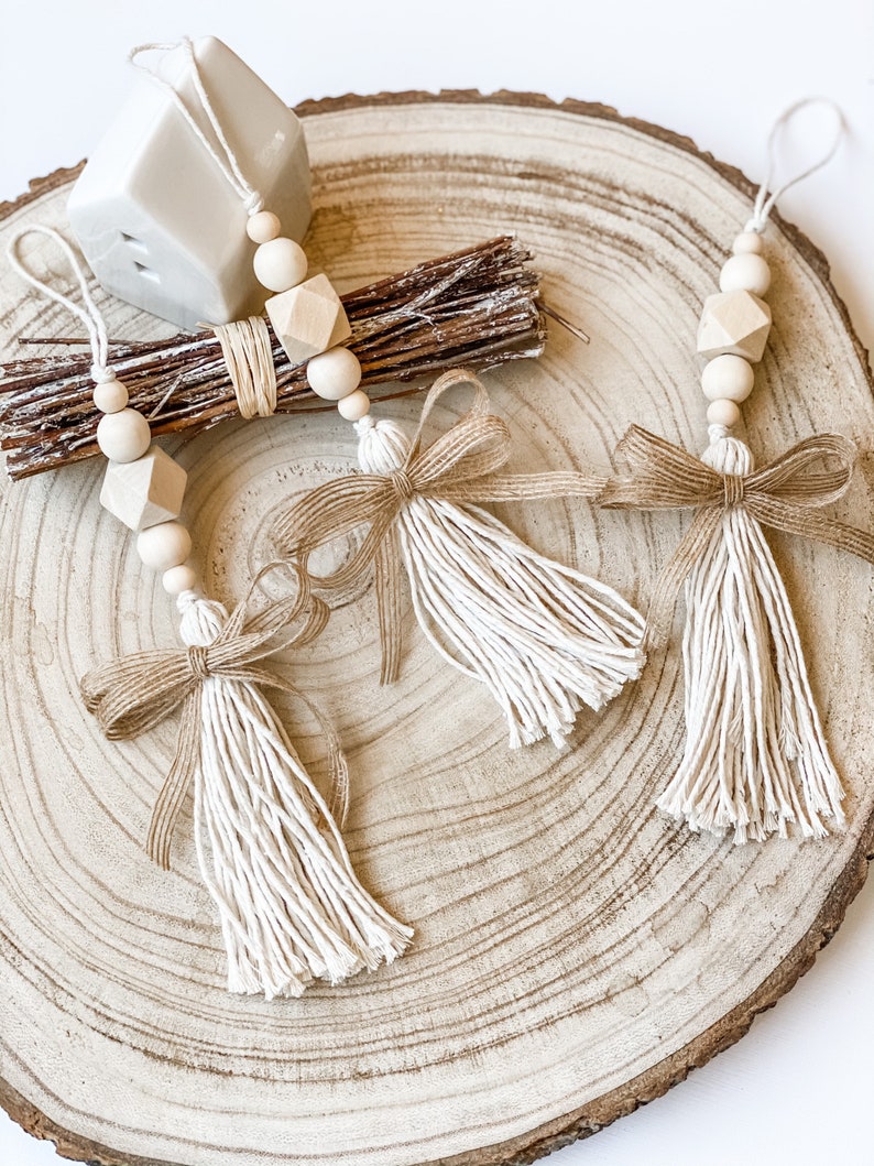May include: Three wooden bead and tassel ornaments with a natural wood and cream colour scheme. The tassels are made of cream coloured yarn and the beads are natural wood. The ornaments are hanging from a wooden slice.