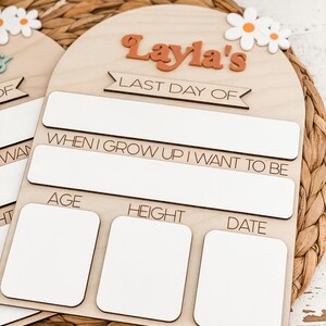 May include: An arched wooden sign featuring the text "Layla's LAST DAY OF" in orange and black, with white daisy accents. It includes blank white rectangular spaces for writing information, with labels for age, height, and date, and a section for career goals.
