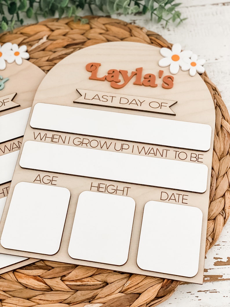 May include: An arched wooden sign featuring the text "Layla's LAST DAY OF" in orange and black, with white daisy accents. It includes blank white rectangular spaces for writing information, with labels for age, height, and date, and a section for career goals.