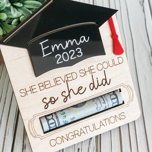 May include: Wooden graduation card with a black graduation cap cutout with the name "Emma 2023" on it. The card has the text "She believed she could so she did" and "Congratulations". A rolled up dollar bill is visible in a slot on the card.