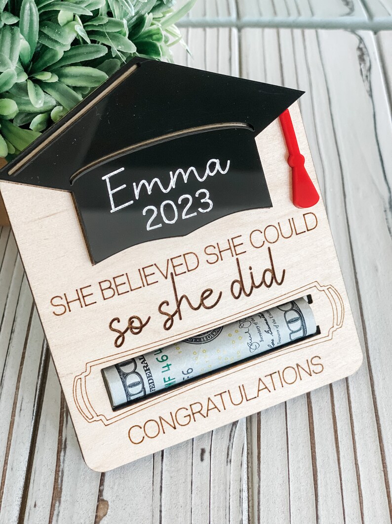 May include: Wooden graduation card with a black graduation cap cutout with the name "Emma 2023" on it. The card has the text "She believed she could so she did" and "Congratulations". A rolled up dollar bill is visible in a slot on the card.