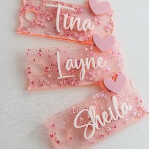 May include: Three pink glitter keychains with white names "Tina", "Layne", and "Sheila" and pink heart charms.