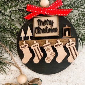 May include: A black and wood Christmas ornament with the words "Merry Christmas" in a square frame. There are five stockings with names on them: Daddy, Mama, Abraham, Abigail, and Adam. The ornament has a red bow with white polka dots.