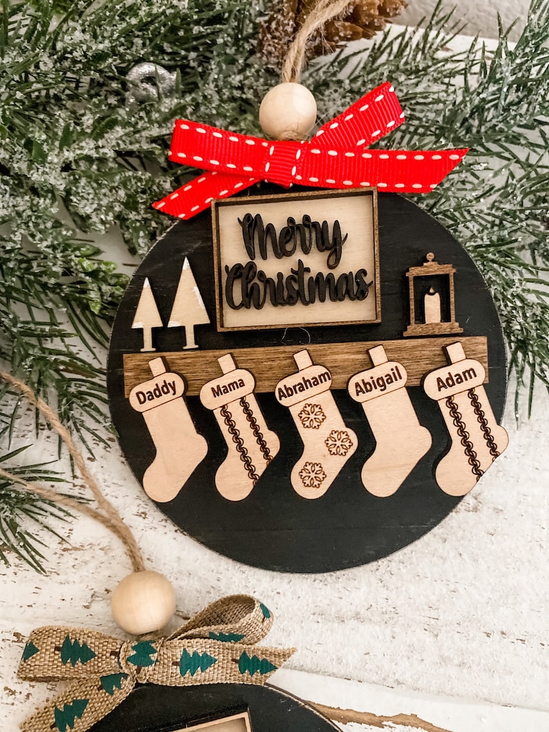 May include: A black and wood Christmas ornament with the words "Merry Christmas" in a square frame. There are five stockings with names on them: Daddy, Mama, Abraham, Abigail, and Adam. The ornament has a red bow with white polka dots.
