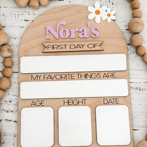 May include: An arched wooden sign featuring the text Nora's FIRST DAY OF, MY FAVORITE THINGS ARE, AGE, HEIGHT, and DATE. The sign includes white rectangular spaces for writing, two white and orange daisy cutouts, and a natural light wood finish.