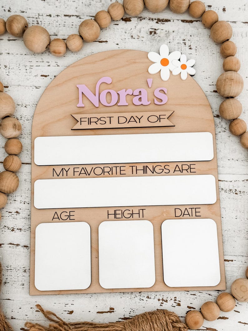 May include: An arched wooden sign featuring the text Nora's FIRST DAY OF, MY FAVORITE THINGS ARE, AGE, HEIGHT, and DATE. The sign includes white rectangular spaces for writing, two white and orange daisy cutouts, and a natural light wood finish.