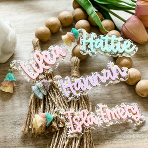 May include: Acrylic name tags with daisy accents and colorful tassels. The names "Lilea", "Hattie", "Hannah", and "Isla Emery" are displayed in a cursive font. The tags are attached to a rope bundle, with wooden beads in the background.
