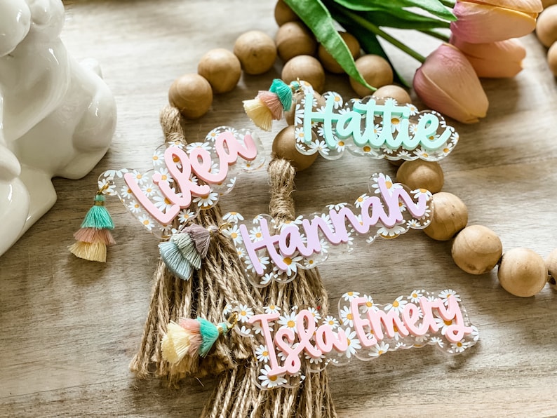 May include: Acrylic name tags with daisy accents and colorful tassels. The names "Lilea", "Hattie", "Hannah", and "Isla Emery" are displayed in a cursive font. The tags are attached to a rope bundle, with wooden beads in the background.