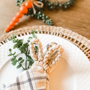 May include: A white crocheted bunny napkin ring with a plaid napkin on a white plate. The napkin ring is a simple design with two loops for the ears.