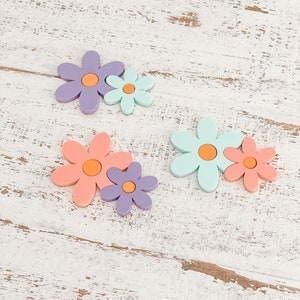 May include: Three pairs of flat, flower-shaped ornaments, each consisting of a larger and a smaller flower joined together. The flowers feature smooth, matte surfaces in pastel shades of purple, mint green, and coral, all with orange centers.