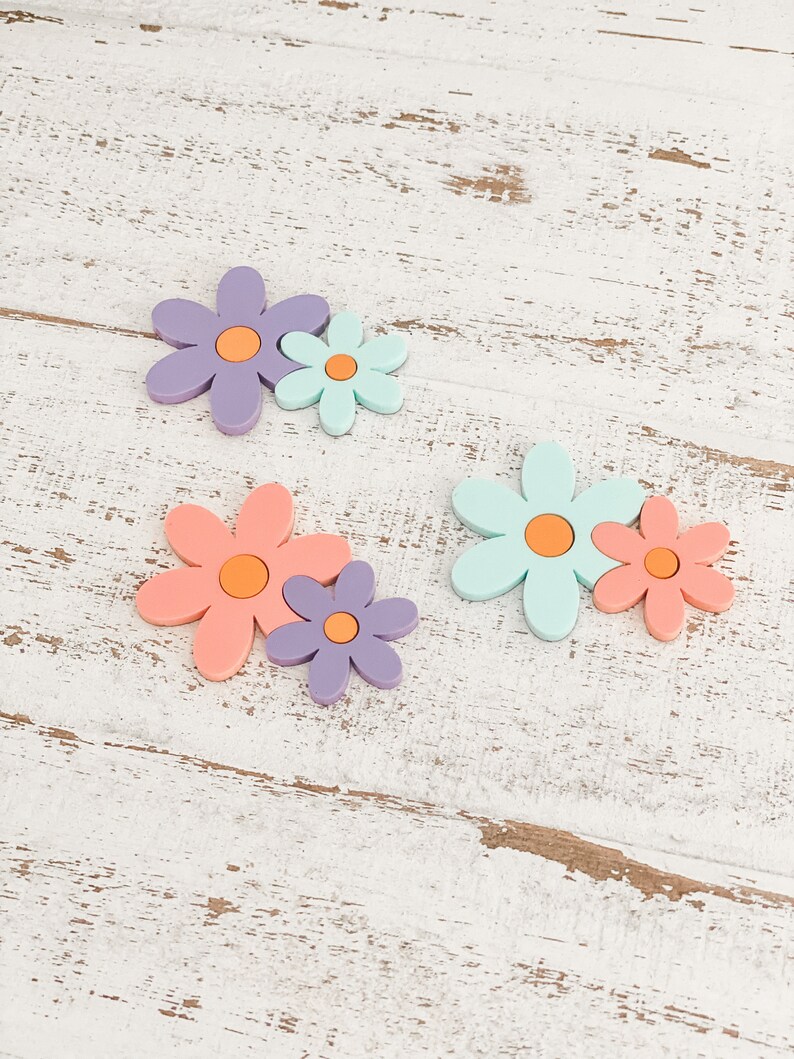 May include: Three pairs of flat, flower-shaped ornaments, each consisting of a larger and a smaller flower joined together. The flowers feature smooth, matte surfaces in pastel shades of purple, mint green, and coral, all with orange centers.