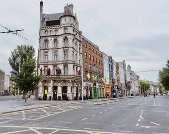 May include: A street scene in Dublin, Ireland, featuring a corner building with ornate architecture. The building has a light-coloured facade with dark accents and statues. Other buildings line the street, and the sky is overcast. A pedestrian crossing and road markings are visible.