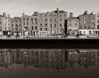 May include: A black and white photograph of a row of brick buildings reflected in a still body of water. The buildings have many windows and are lined up along the water's edge.