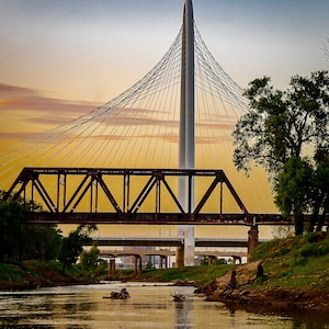 May include: A large, modern cable-stayed bridge with a white tower and a steel truss bridge underneath. The bridge is over a river with green banks and a blue sky with clouds in the background.