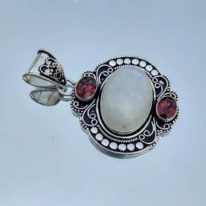 Puede incluir: Un colgante de plata con una gran piedra de luna ovalada y dos piedras de granate rojo m&aacute;s peque&ntilde;as. El colgante tiene un dise&ntilde;o intrincado con espirales y puntos.