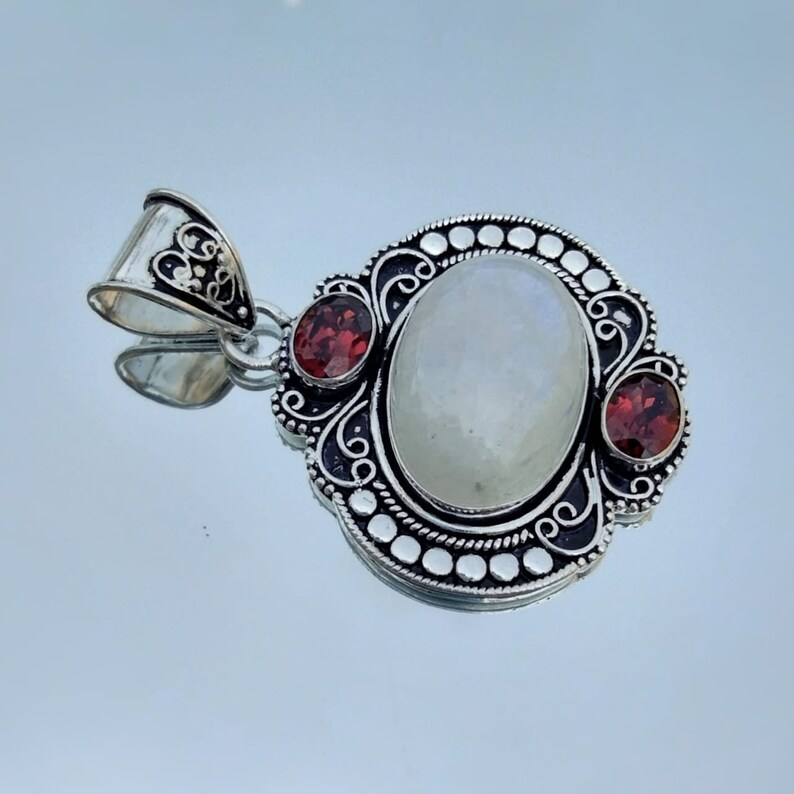 Puede incluir: Un colgante de plata con una gran piedra de luna ovalada y dos piedras de granate rojo m&aacute;s peque&ntilde;as. El colgante tiene un dise&ntilde;o intrincado con espirales y puntos.