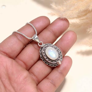 May include: A silver-toned pendant necklace featuring an oval, iridescent moonstone. The moonstone is set within an ornate, filigree-style frame. The chain is a delicate, twisted design, and the pendant is held in a hand.