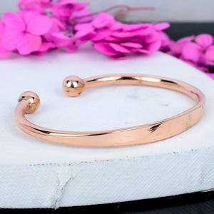May include: A rose gold cuff bracelet with rounded ball ends. The open design features a smooth, polished surface. The bracelet is displayed on a white surface with bright pink flowers in the background. A simple, elegant piece of jewelry.