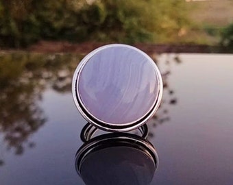 Puede incluir: Un anillo de plata con una piedra de ágata grande, redonda y de color púrpura claro. La piedra tiene un patrón de remolino.
