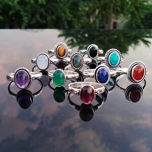 May include: A collection of sterling silver rings with various oval-shaped gemstones, including amethyst, onyx, and turquoise. The rings are displayed on a reflective surface, showcasing their polished metal and colorful stones. The gemstones are set in a bezel setting.