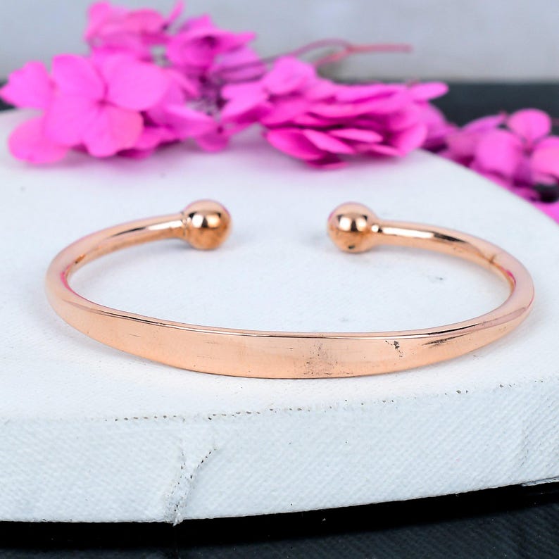 May include: A rose gold cuff bracelet with rounded ball ends. The open design features a smooth, polished surface. The bracelet is displayed on a white surface with pink flowers in the background. Jewelry.