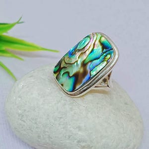 May include: A rectangular ring featuring an iridescent abalone shell. The shell displays a mix of blue, green, and brown hues, set in a silver-toned metal band. The ring is displayed on a white stone.