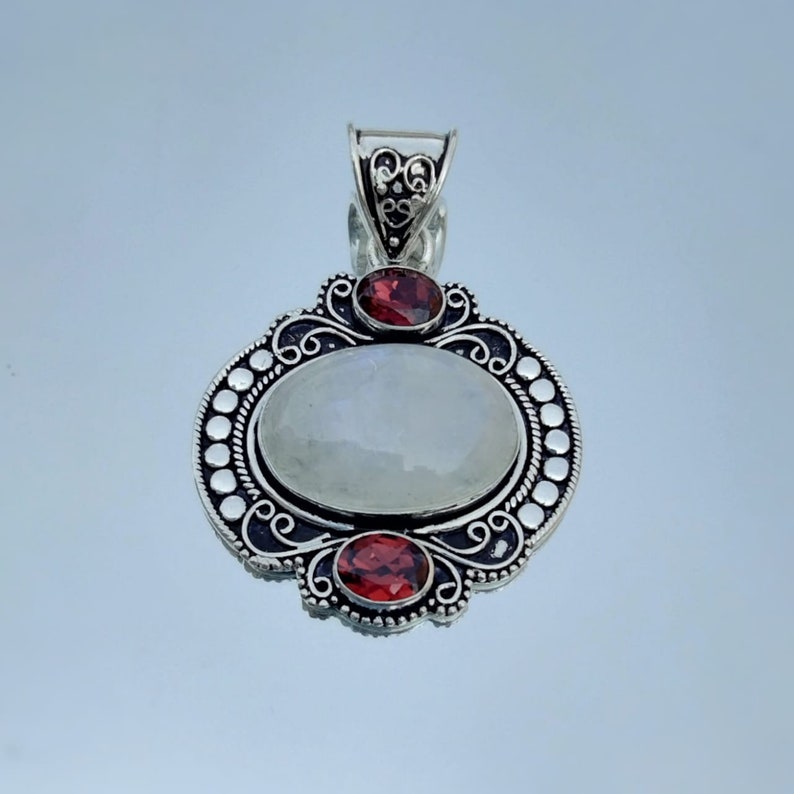 Puede incluir: Un colgante de plata con una piedra de luna ovalada y dos acentos de granate rojo. El colgante tiene un dise&ntilde;o filigrana detallado.