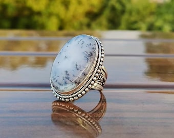 Puede incluir: Anillo de plata con una piedra preciosa ovalada blanca con inclusiones dendríticas negras. El anillo tiene un borde detallado con cuentas y detalles ornamentados en la banda. La piedra preciosa mide aproximadamente 3,8 cm de largo.