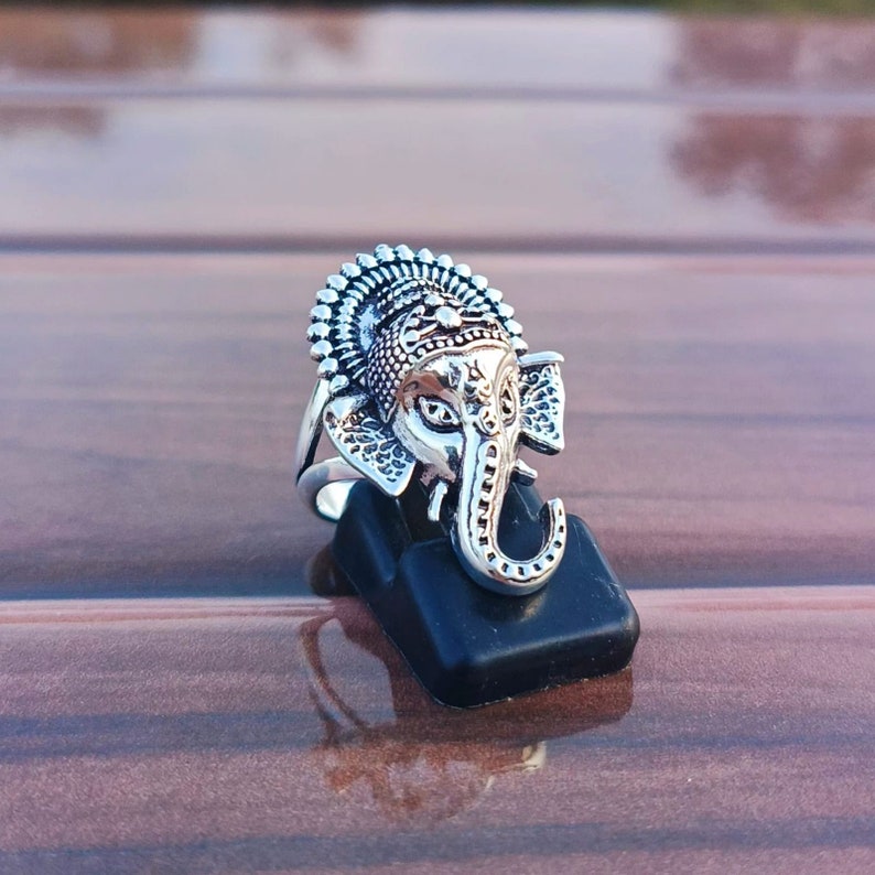 K&ouml;nnte beinhalten: Silberring mit einem detaillierten Elefantenkopfdesign mit komplizierten Mustern. Der R&uuml;ssel des Elefanten ist nach oben gerollt, und der Ring wird auf einem schwarzen Ringst&auml;nder pr&auml;sentiert.